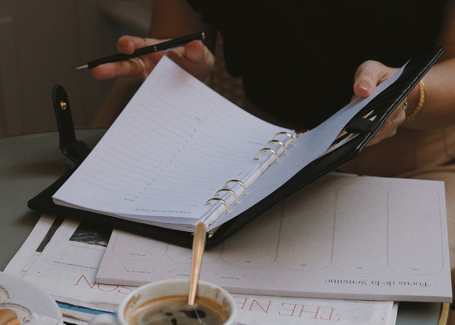 Femme qui travaille en terrasse, elle écrit dans un agenda à anneaux, il y a un semainier et un journal sur la table