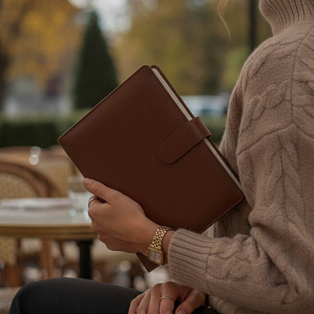 femme en terrasse, pull en maille marron, elle tient un agenda qu'elle a composé elle même, elle porte des bijoux et une montre dorée